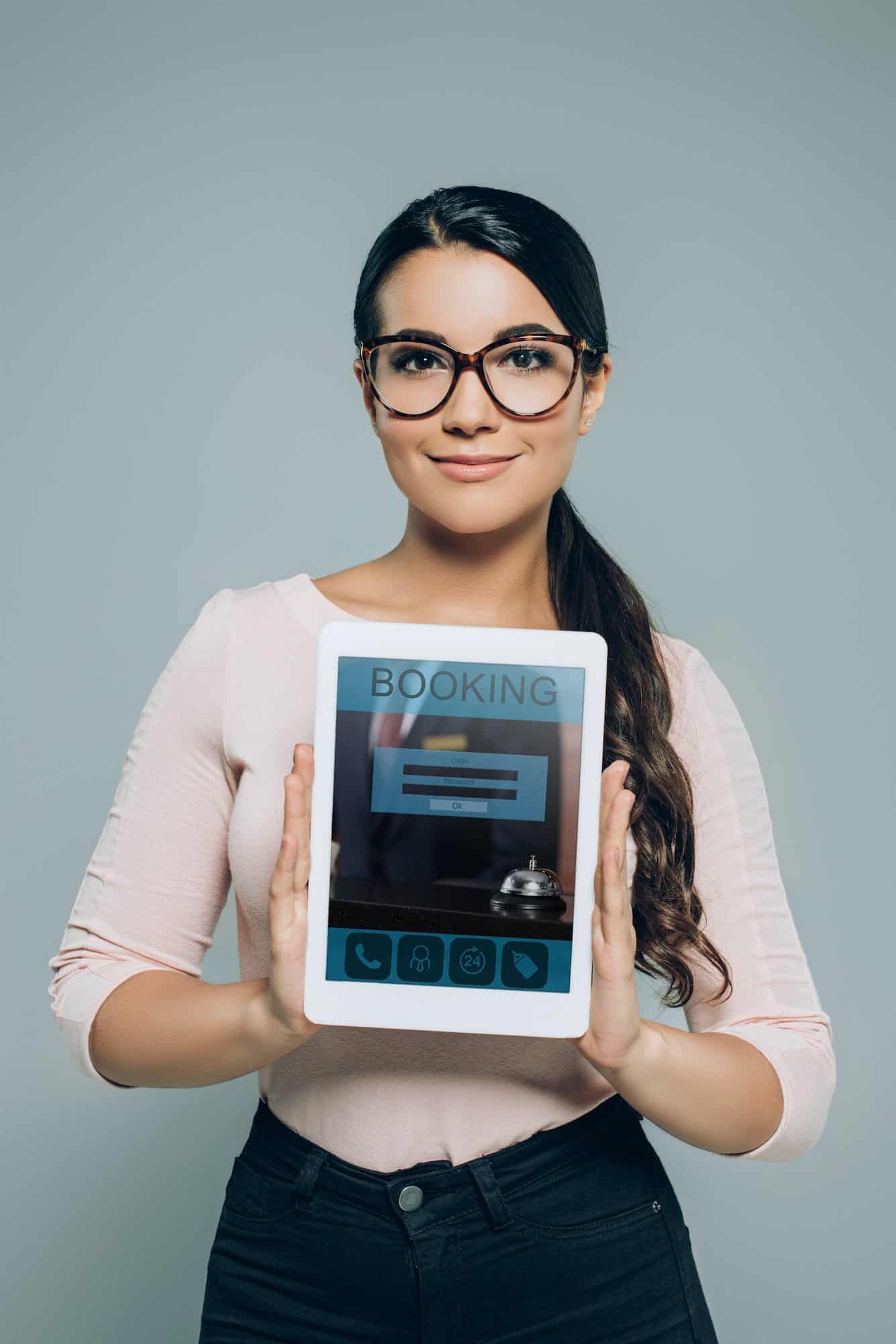 portrait of smiling brunette woman in eyeglasses showing tablet with booking website on screen in